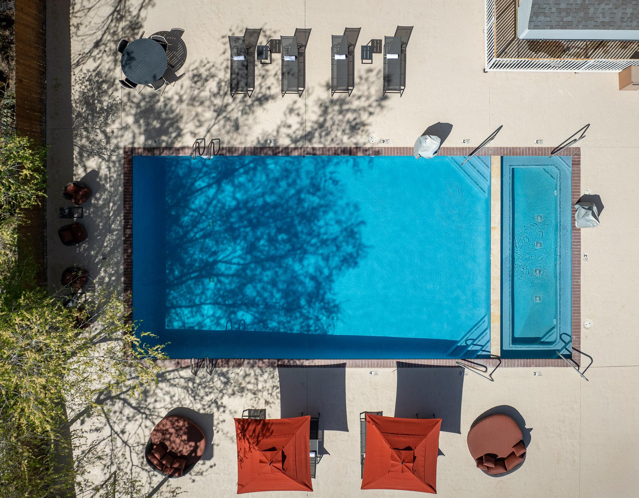 Aerial view of a rectangular pool with a small hot tub and red brick border, surrounded by orange chairs and trees casting shadows.